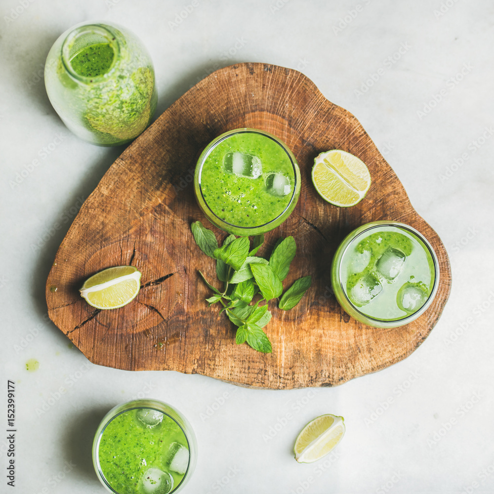 Fresh green smoothie in bottle and glasses with ice cubes, mint and lime on wooden board over grey m