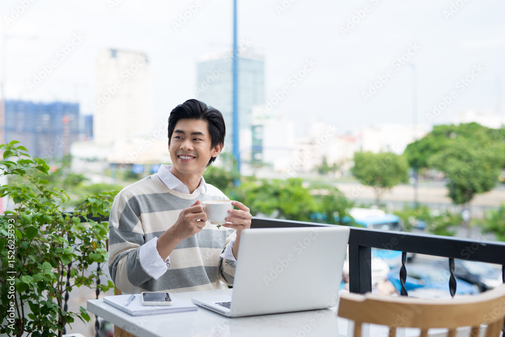 Handsome asian young man working on laptop and smiling while enjoying coffee in cafe