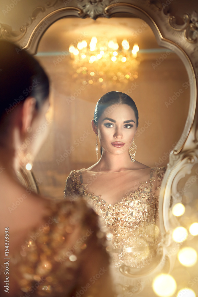 Beauty glamour lady looking in the mirror. Gorgeous woman in beautiful evening dress in luxurious st