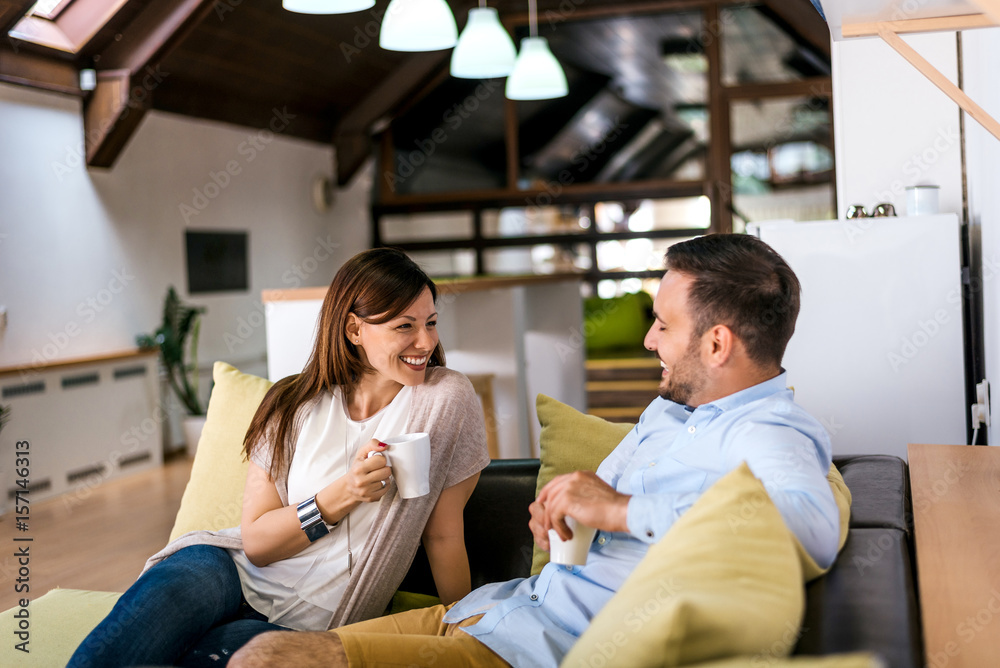 Beautiful young couple drink coffee while sitting on a sofa, looking each other