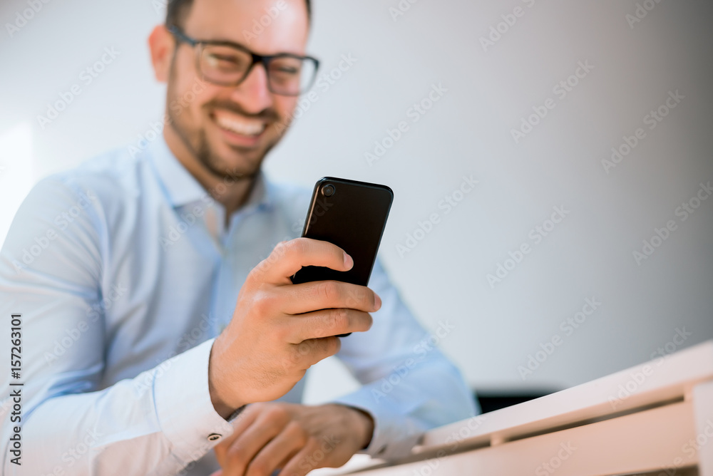 Smiling young businessman in glasses using his martphone at workplce.