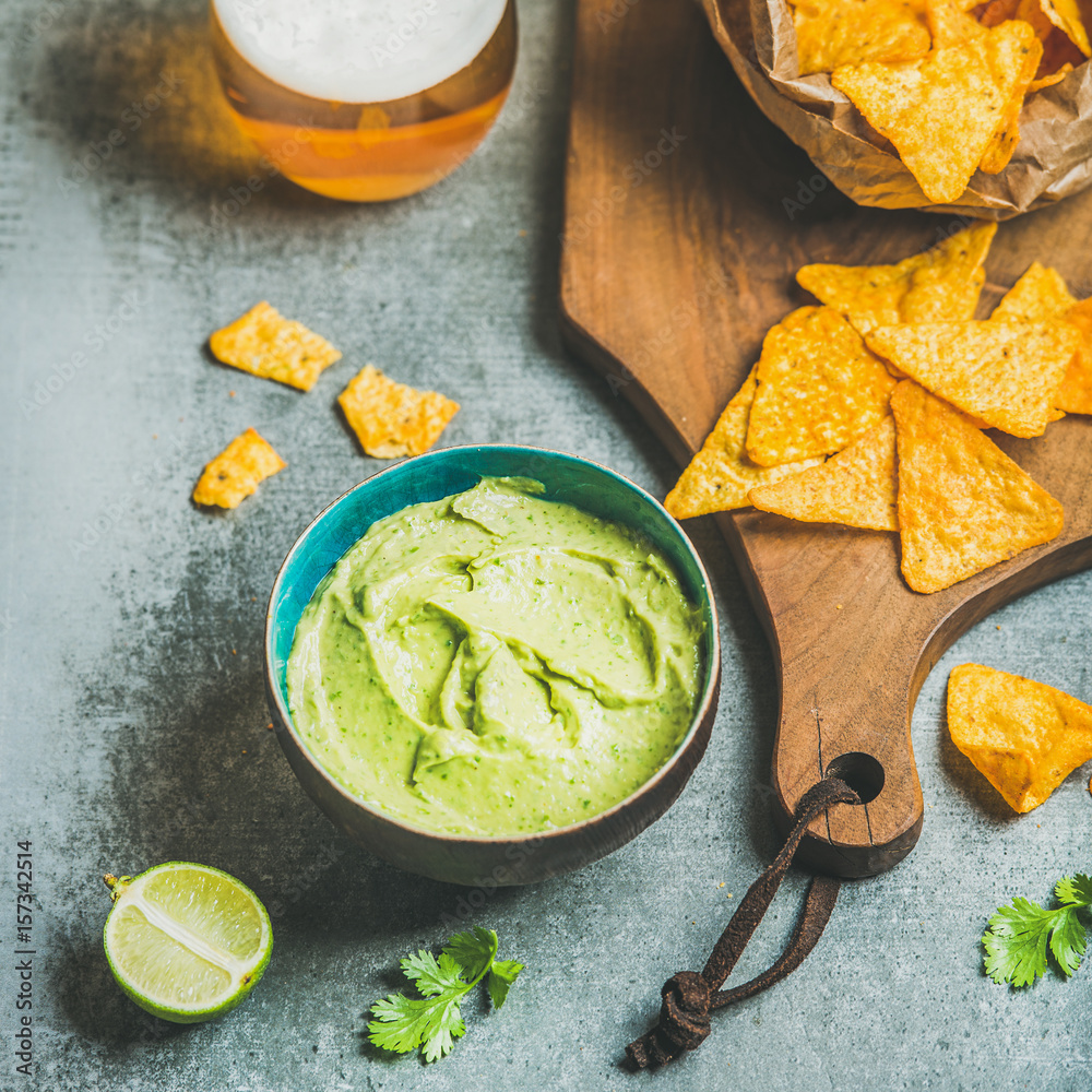 Mexican corn chips, fresh guacamole sauce and glass of beer on wooden serving board over grey concre