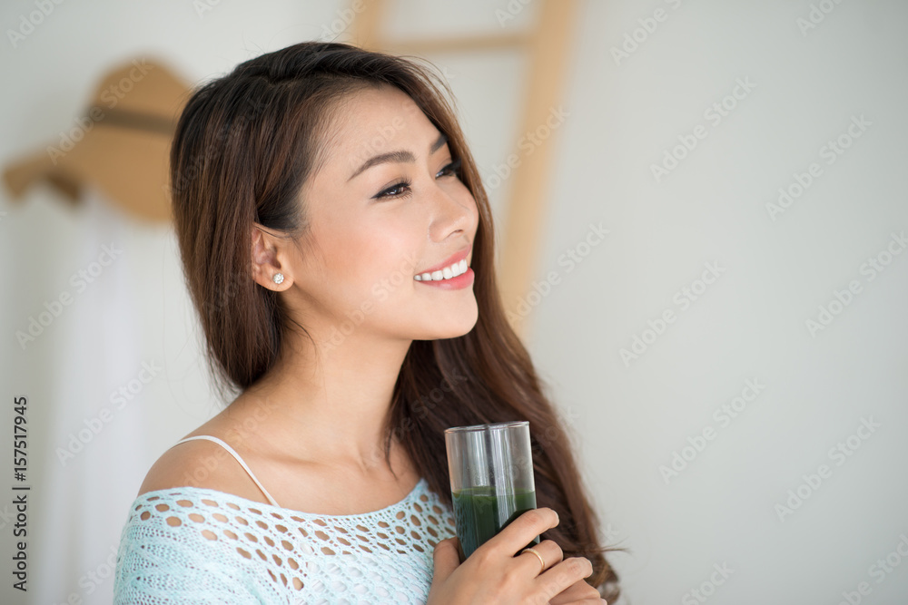 Pretty young asian woman drinking green fresh vegetable juice or smoothie from glass at home