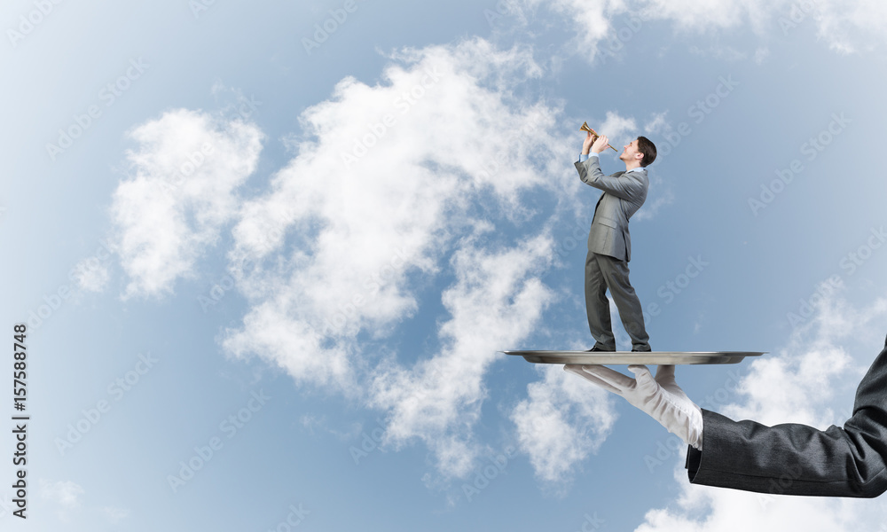 Businessman on metal tray playing fife against blue sky background