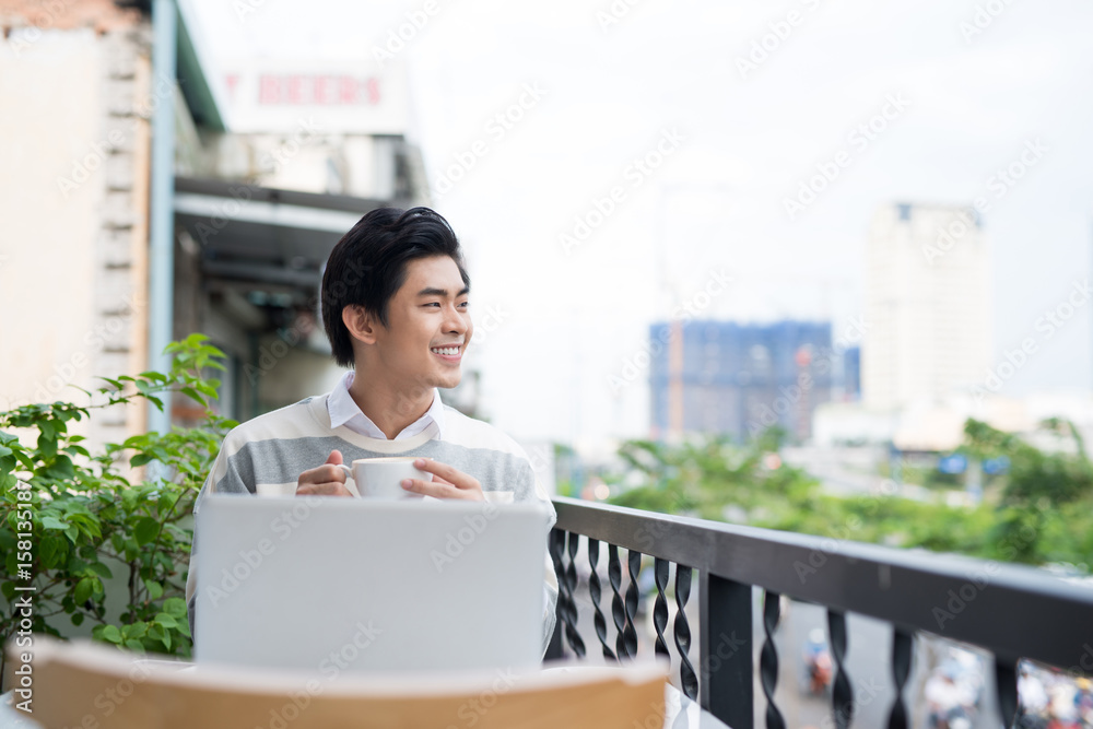 Handsome asian young man working on laptop and smiling while enjoying coffee looking out at the view