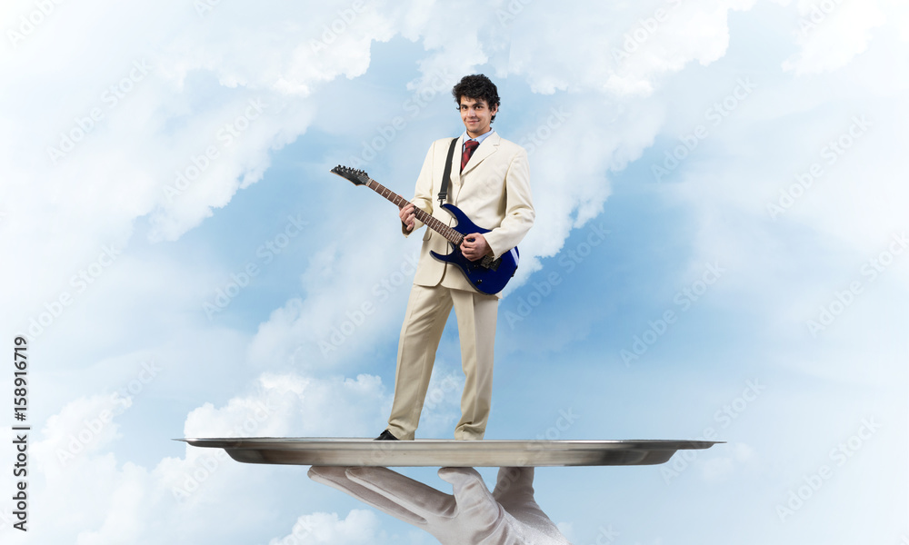 Businessman on metal tray playing electric guitar against blue sky background