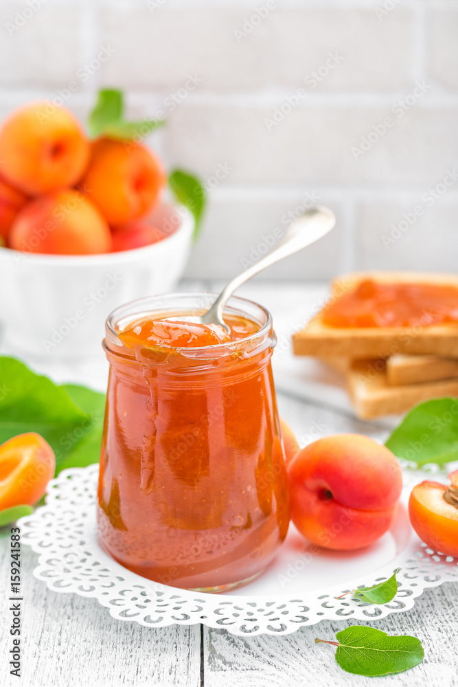 Apricot jam in a jar and fresh fruits with leaves on white wooden table, breakfast