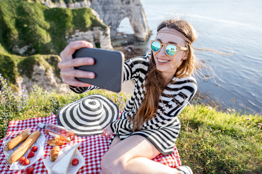 Young woman making selfie portrait with phone during the picnic outdoors sitting on the beautiful ro