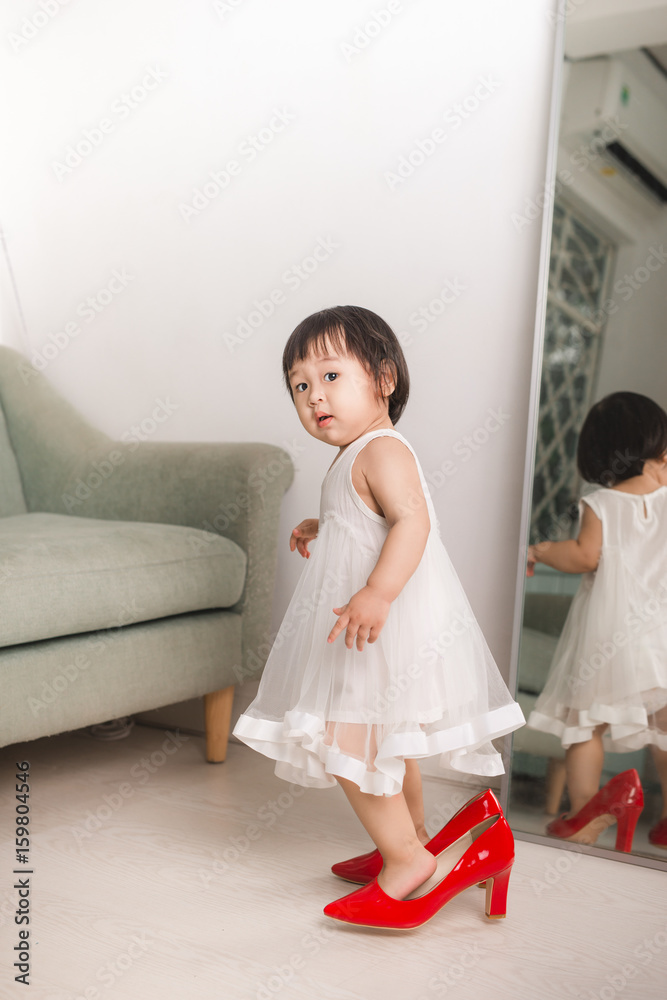 Cute funny little baby girl walks at home trying moms red high heel shoes