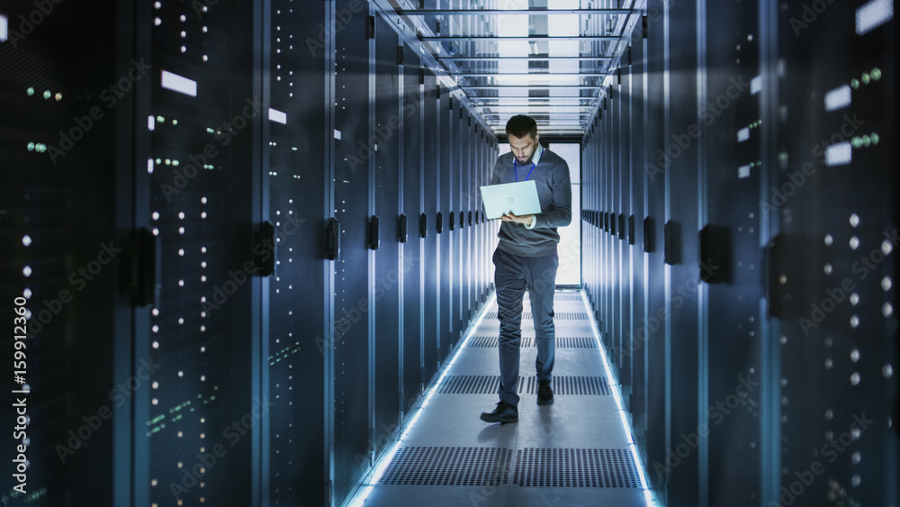 IT Technician Works on Laptop in Big Data Center full of Rack Servers. He Runs Diagnostics and Maint