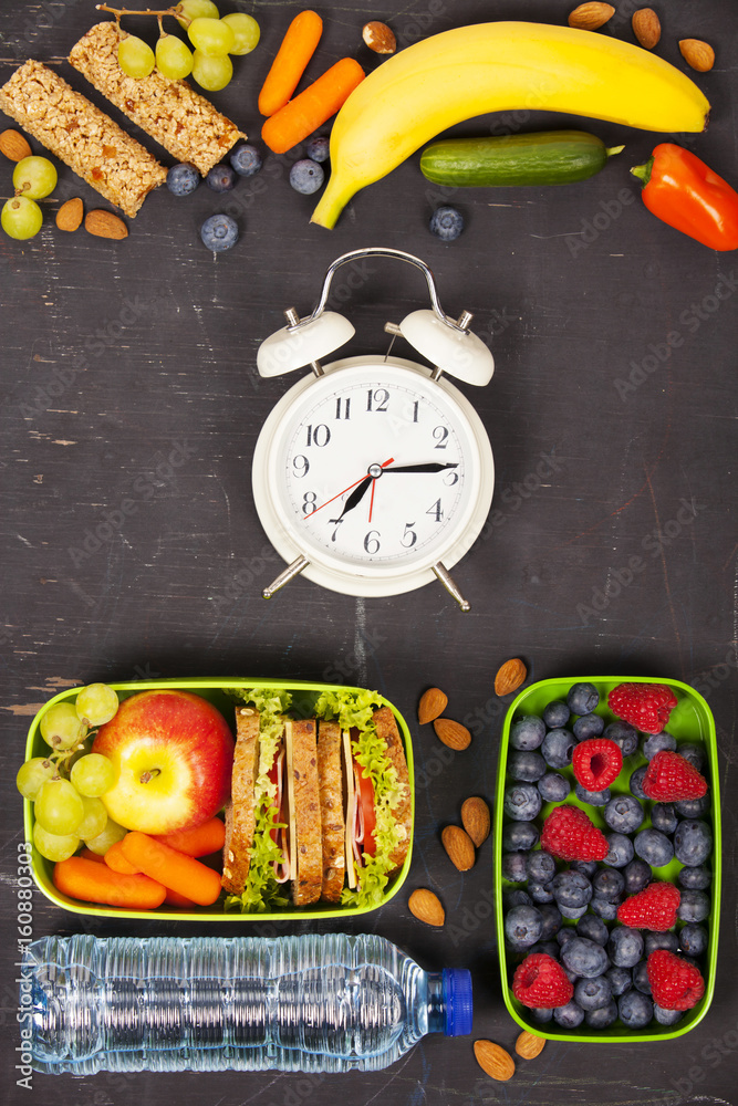 Sandwich, apple, grape, carrot, berry in plastic lunch boxes, alarm clock and bottle of water