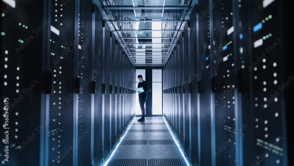 Male IT Engineer Works on a Laptop in a Big Data Center. Rows of Rack Servers are Seen.