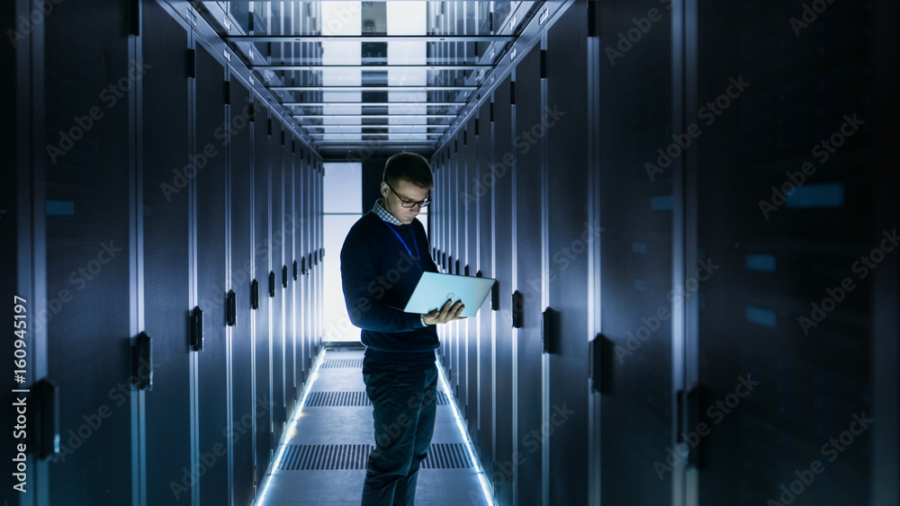 Male IT Engineer Works on a Laptop in front of Server Cabinet at a Big Data Center. Rows of Rack Ser