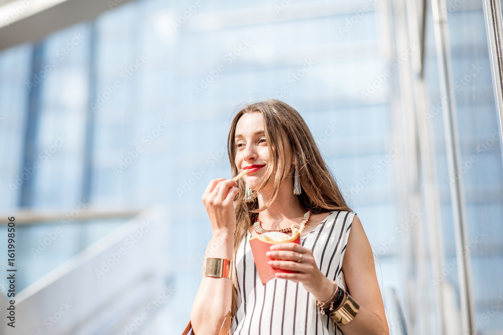 Young businesswoman having a quick snack with frech fries outdoors on the stairs near the modern off