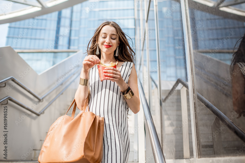 Young businesswoman having a quick snack with frech fries outdoors on the stairs near the modern off