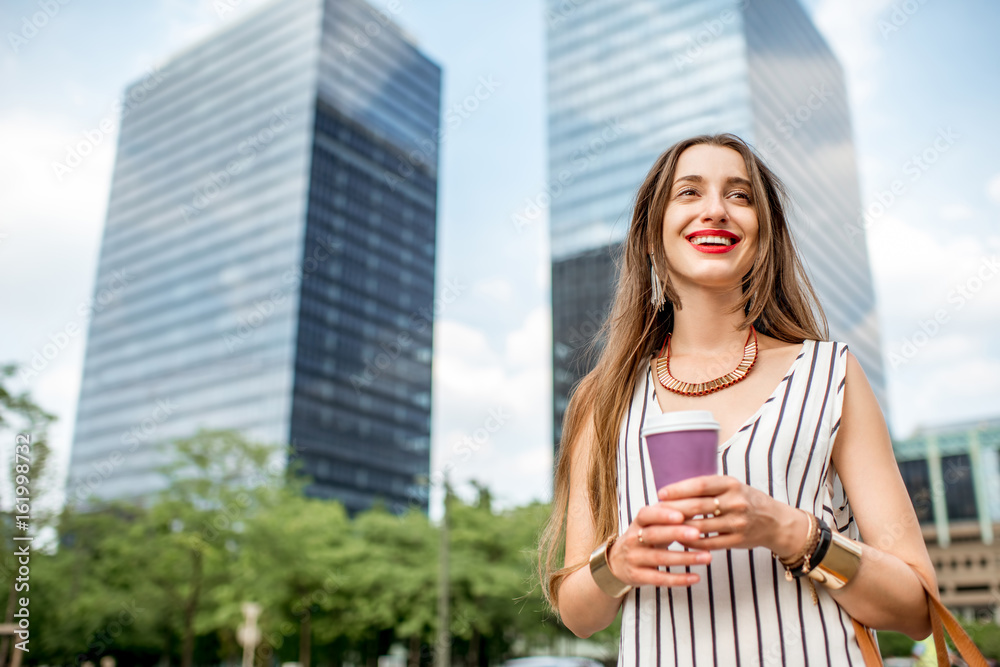 Young and smiling businesswoman having a coffee break standing outdoors on the skyscrapers backgroun