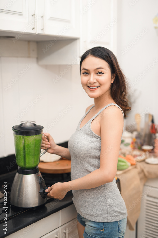 微笑的亚洲女人在家里厨房的搅拌机里用新鲜蔬菜做奶昔。