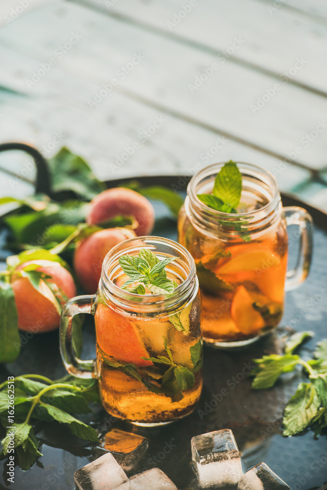 Summer refreshing cold peach ice tea with fresh mint in glass jars on metal tray over rustic wooden 