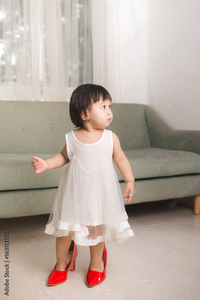 Cute funny little baby girl walks at home trying moms red high heel shoes