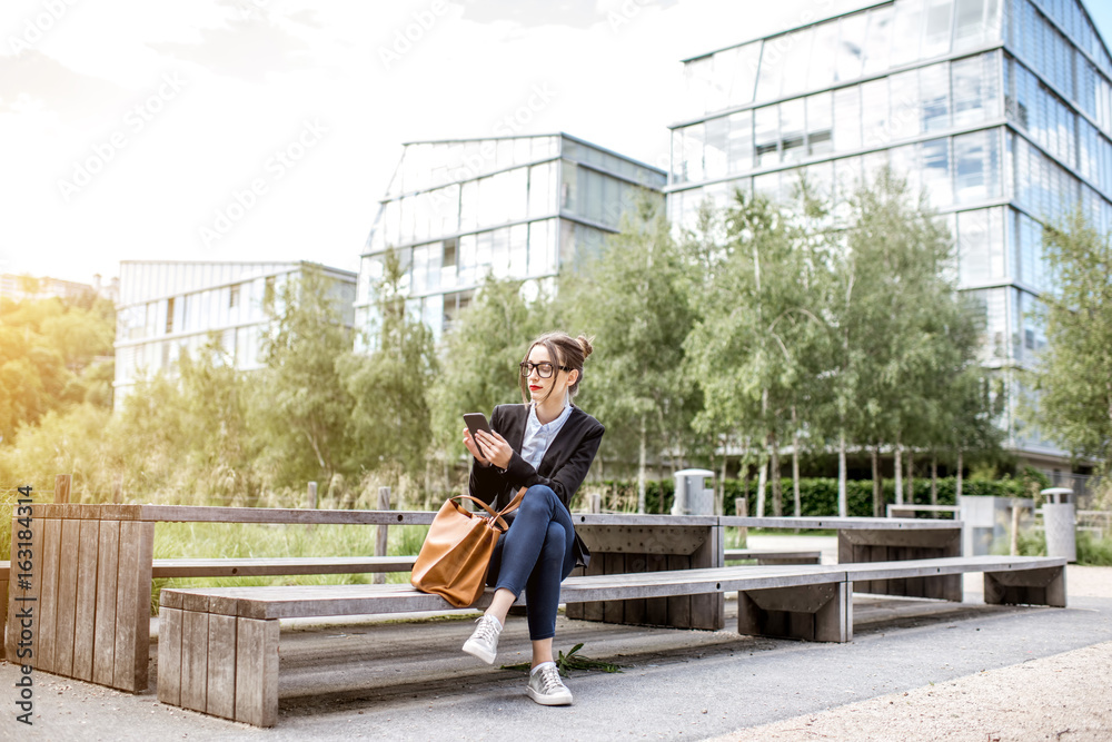 Young businesswoman sitting on the bench with phone outdoors at the modern office district