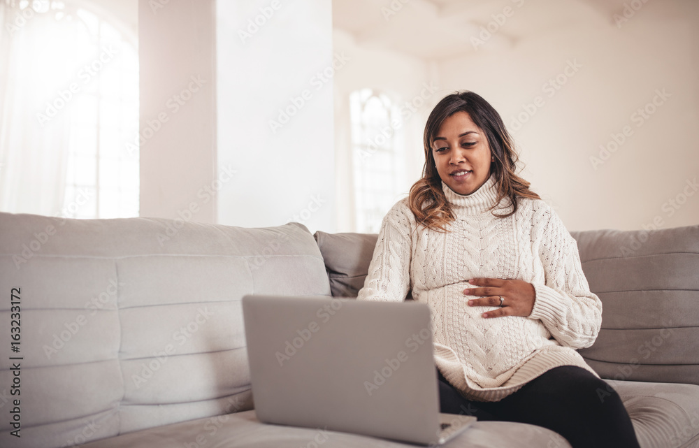 Beautiful pregnant woman with laptop sitting on sofa