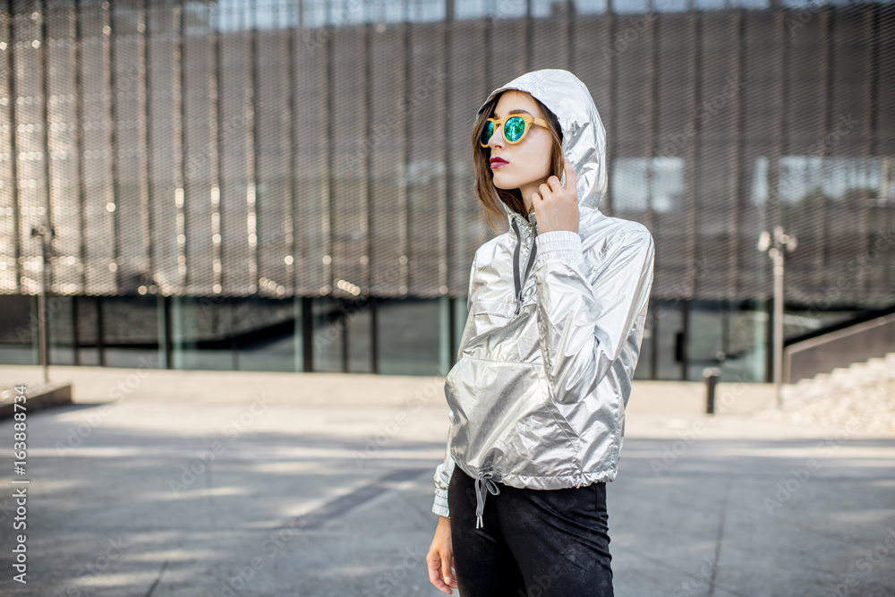 Lifestyle portrait of a stylish woman in silver jacket standing outdoors in the modern urban environ