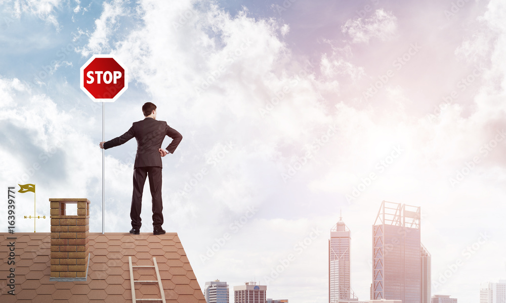 Caucasian businessman on brick house roof showing stop road sign. Mixed media