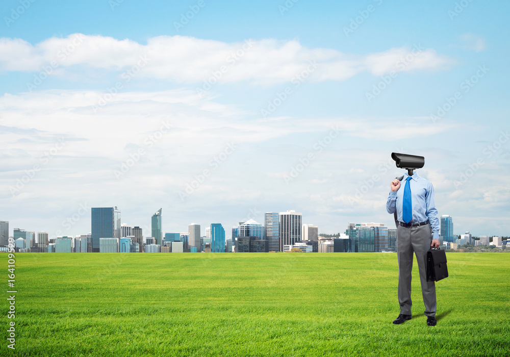 Camera headed man standing on green grass against modern cityscape