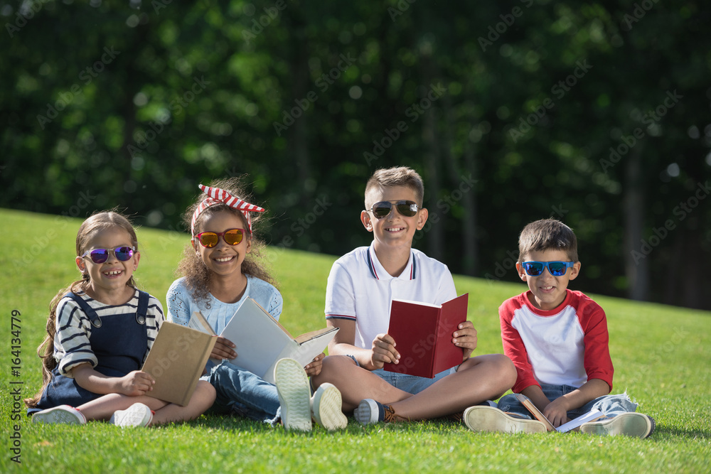 Cute smiling multiethnic kids in sunglasses holding books and smiling at camera in park