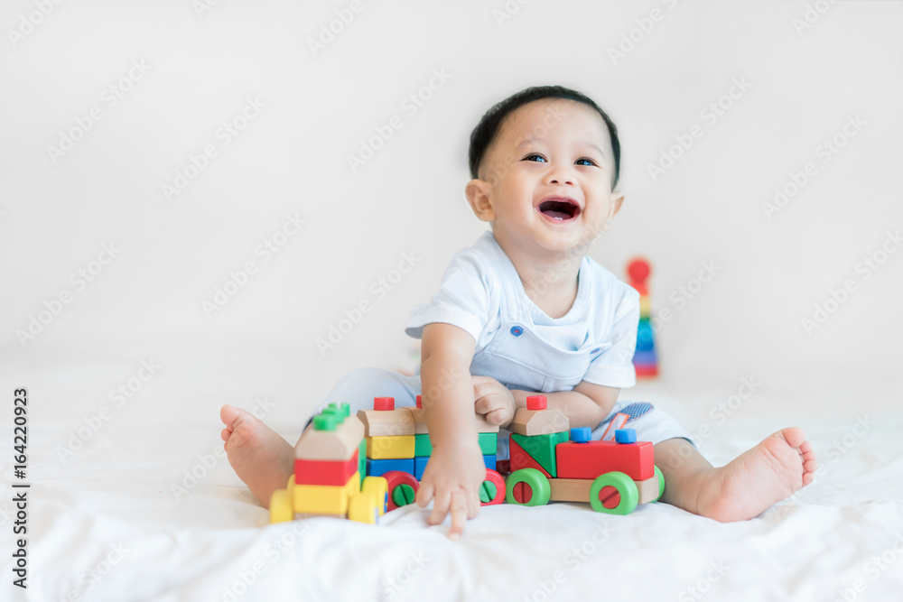 Adorable Asian baby boy 9 months sitting on bed and playing with color wooden train toys at home..