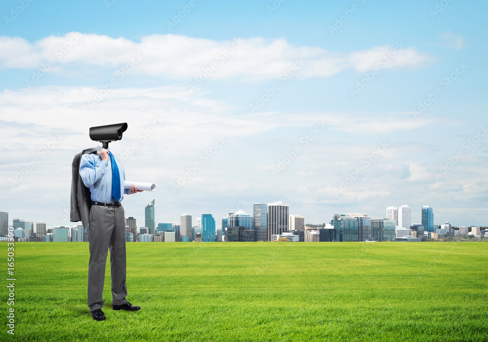 Camera headed man standing on green grass against modern cityscape