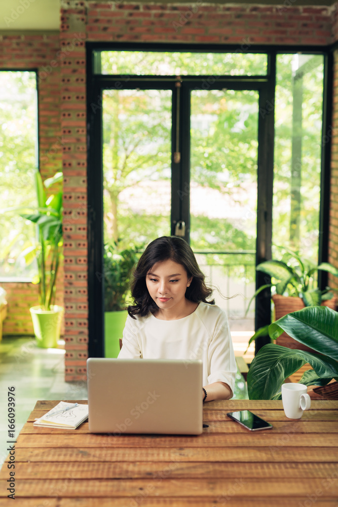 Beautiful young smiling woman working on laptop while sitting on bed at home near big window.