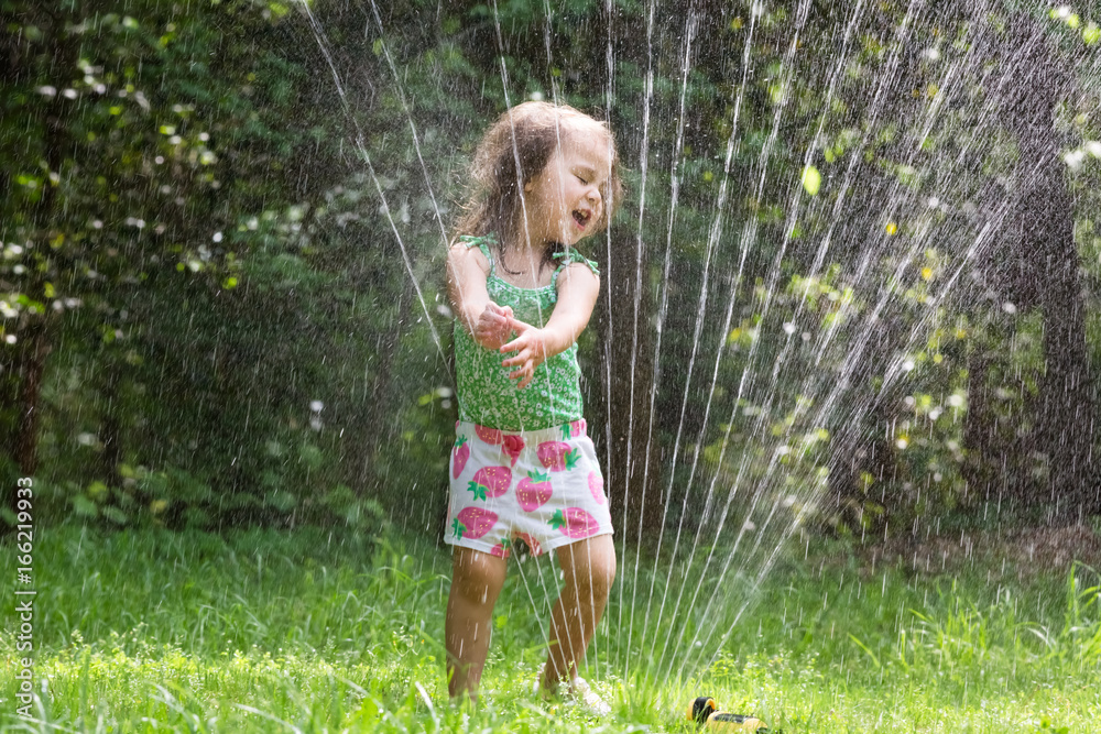 Happy toddler girl playing in a sprinkler on a hot summer day