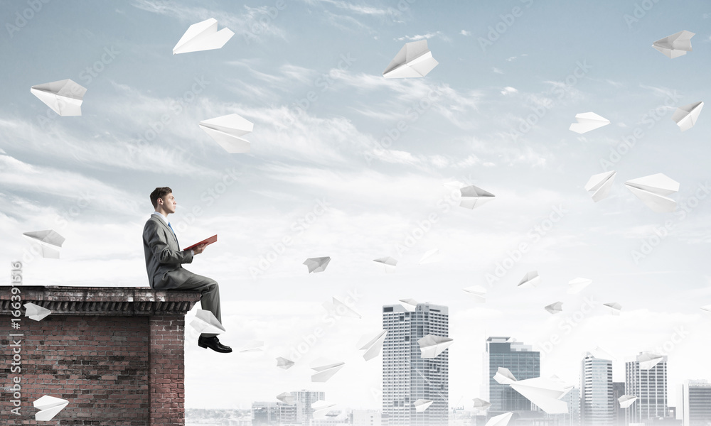 Man on roof edge reading book and cityscape at background