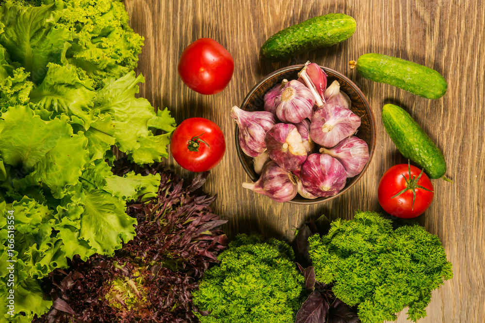 A full bowl with garlic bulbs. Tomatoes and cucumbers on a wooden table. Green salad in the backgrou