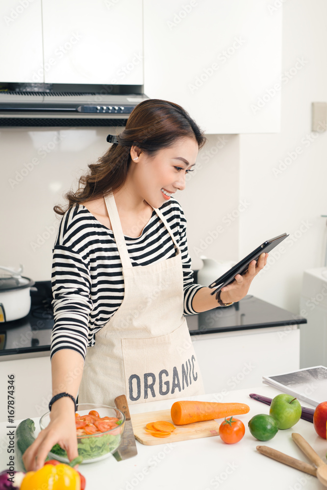 美丽的亚洲女人根据平板电脑屏幕上的食谱烹饪