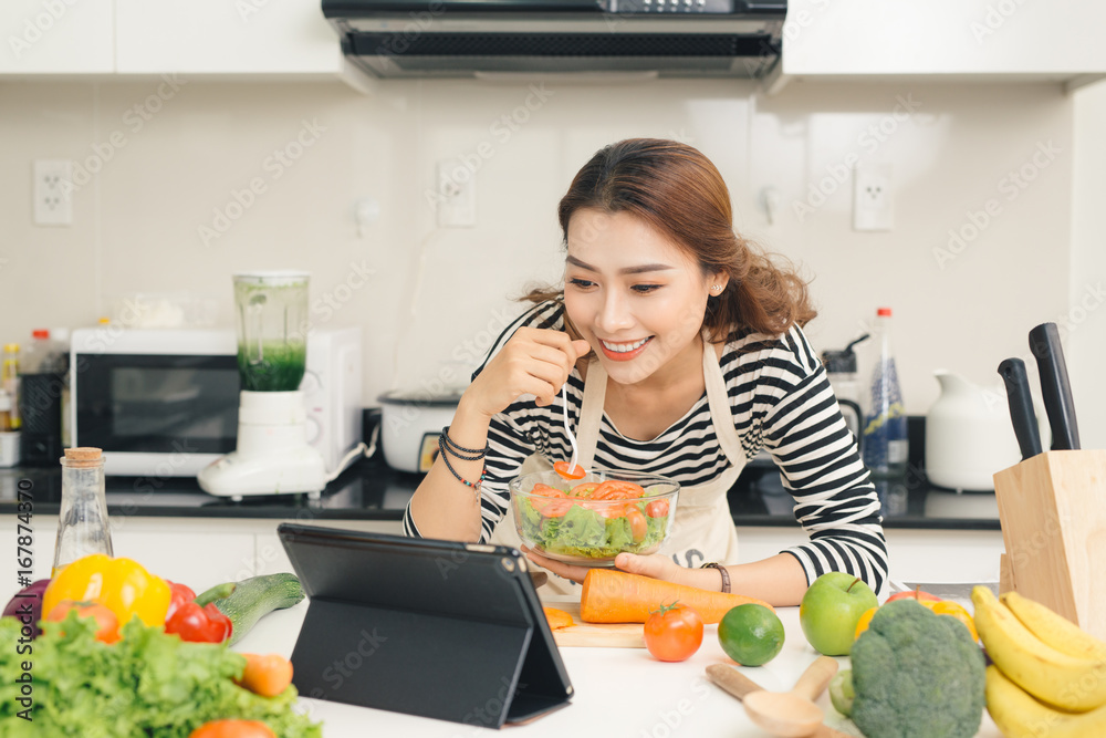 微笑的越南女人在数字平板电脑上看电视节目，在家里厨房做饭