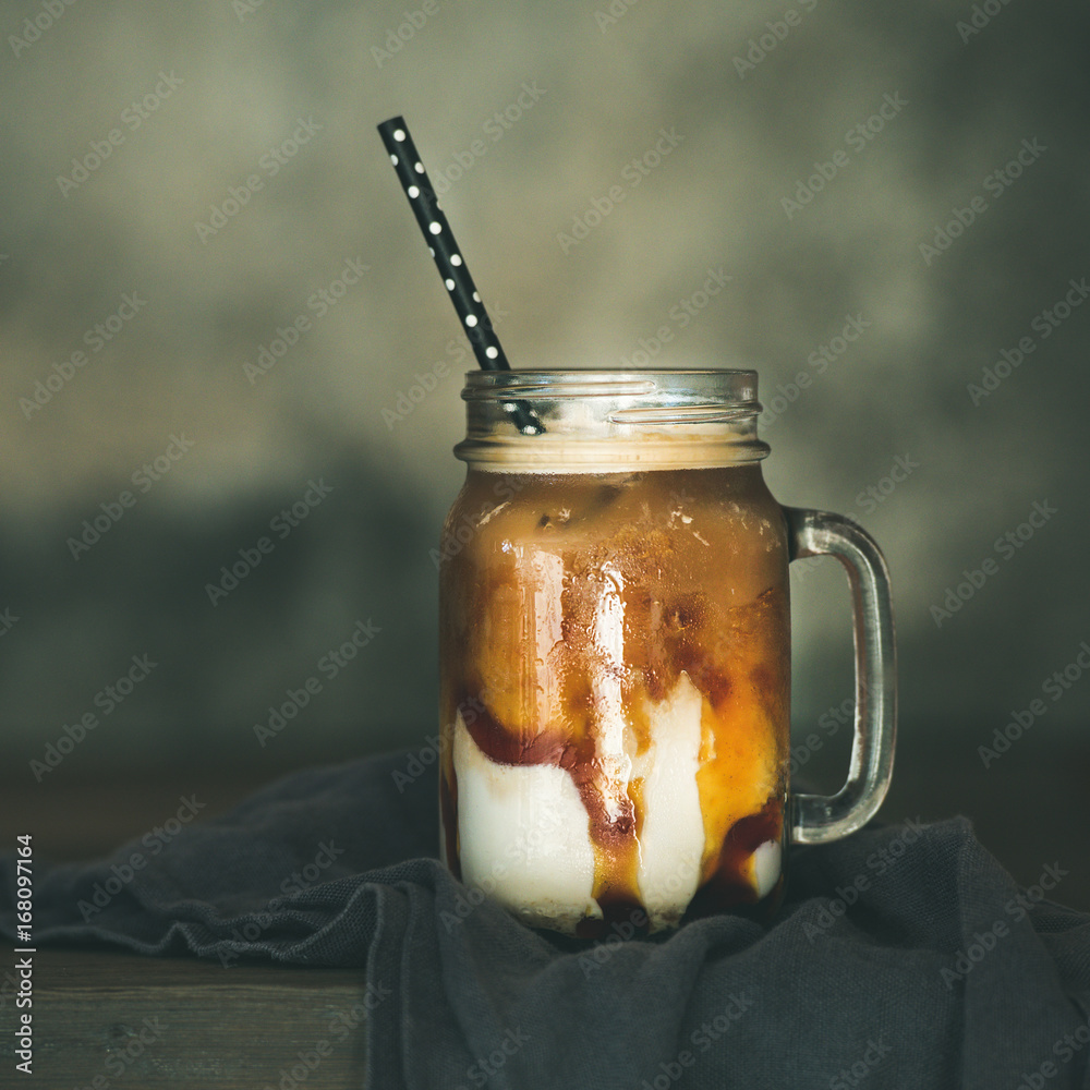 Iced caramel macciato coffee with milk in glass jar with straw on dark rustic wooden table, selectiv