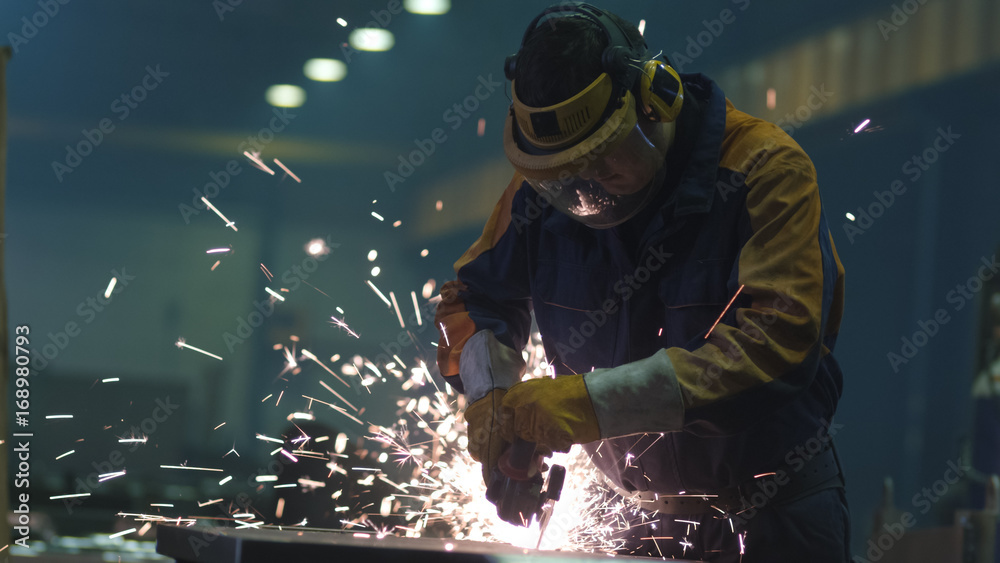 Heavy industry worker at a factory is working with metal on a angle grinder while hot sparks are pro