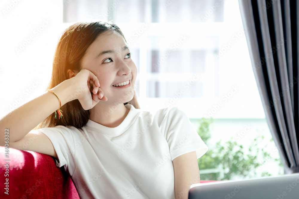 Asian teenage girl using a laptop computer to check her orders online, and relax on a red sofa in he