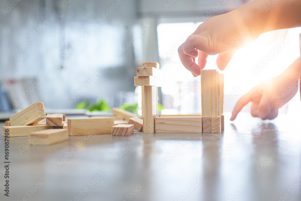 Hand of kid playing a blocks wood tower game of architectural project with sun flare and blur backgr