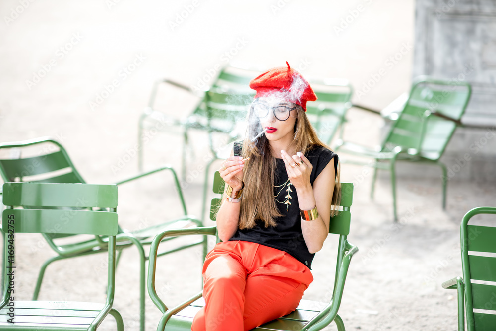 Young stylish woman in red cap smoking a cigarette sitting outdoors on the green chairs in Paris