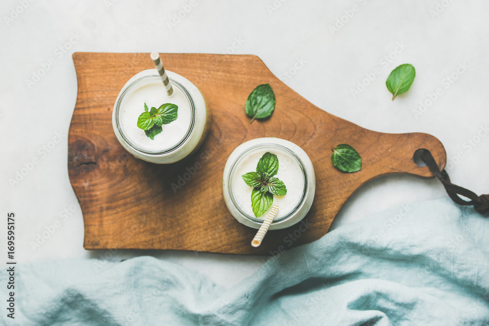 Ombre layered green smoothies with mint in glass jars with straws on wooden board over light grey ba