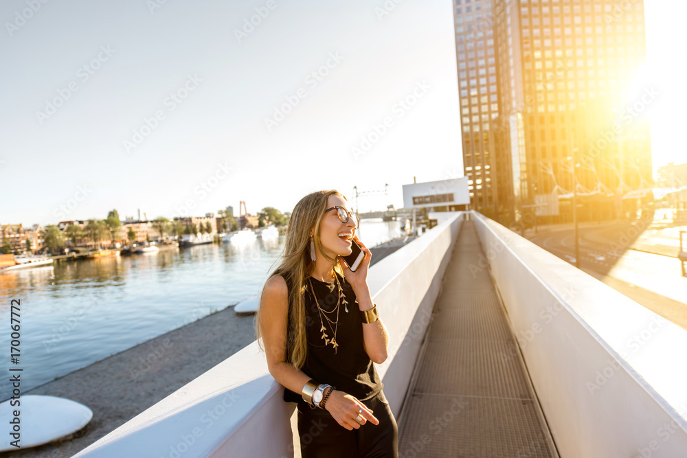 Lifestyle portrait of a stylish woman standing with phone on the modern bridge with skyscrapers on t