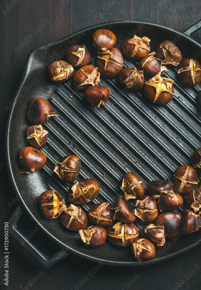 Roasted chestnuts in cast iron grilling pan over dark scorched wooden background, top view, copy spa