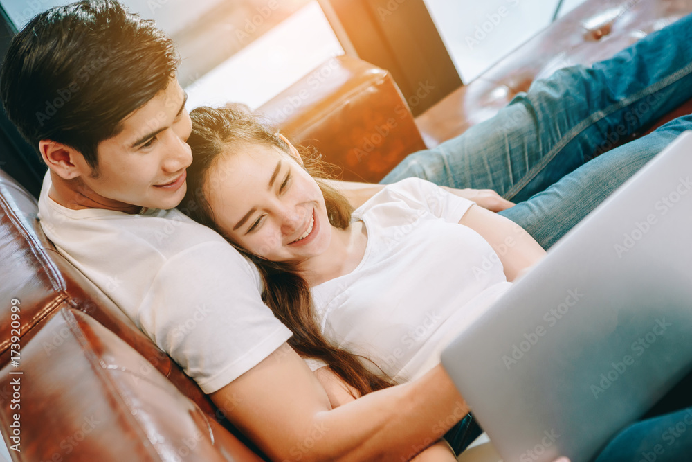 Teen Asian couple looking at laptop computer use on leather sofas in their beautiful home.