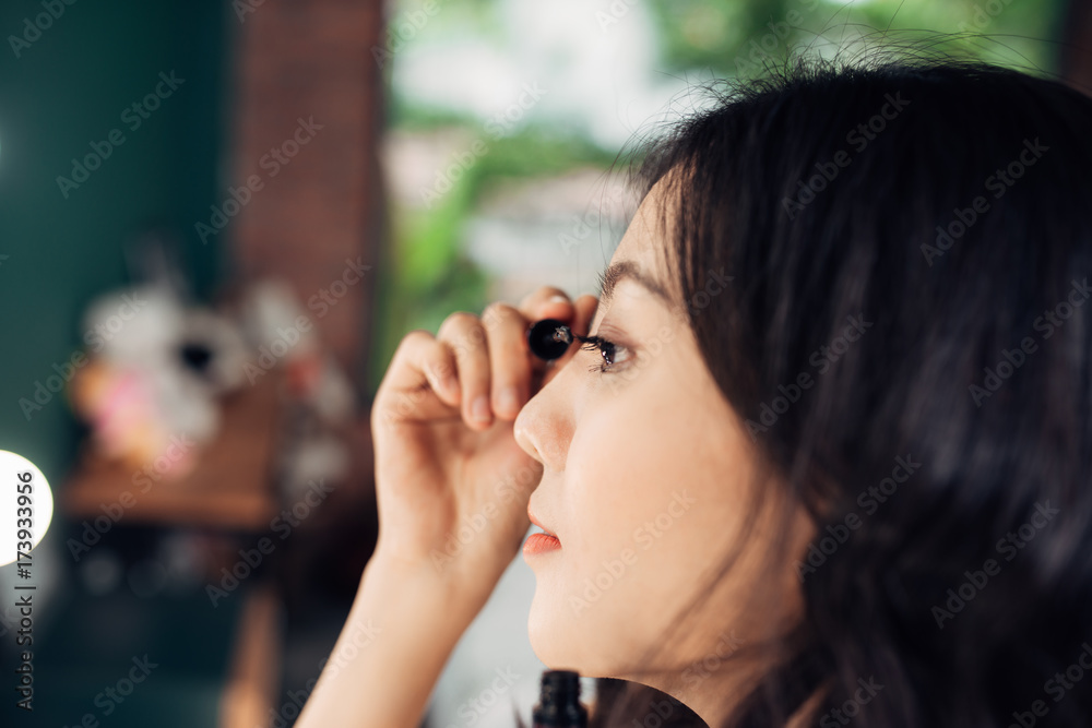 Portrait of beautiful young woman looking at the mirror applying black mascara on eyelashes at home