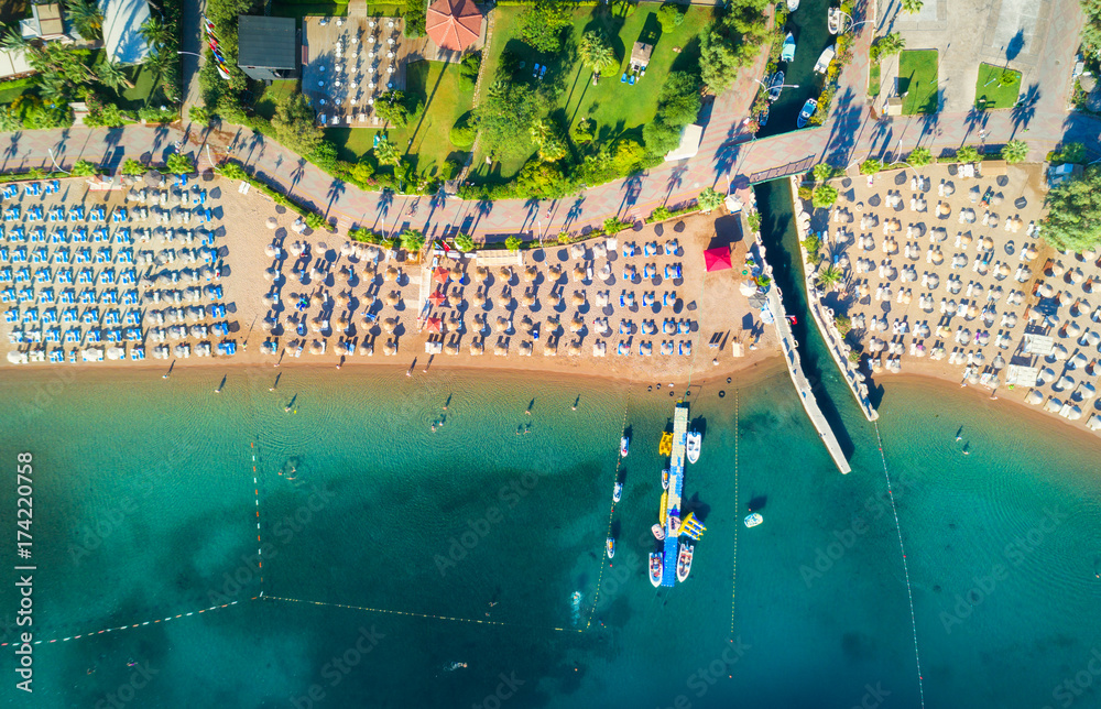 Aerial view of transparent turquoise sea, beautiful sandy beach with colorful chaise-lounges, boats,