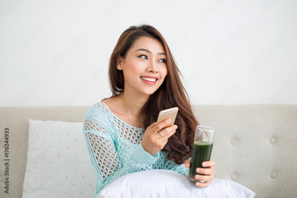 Smiling young asian woman drinking green fresh vegetable juice or smoothie from glass using smartpho