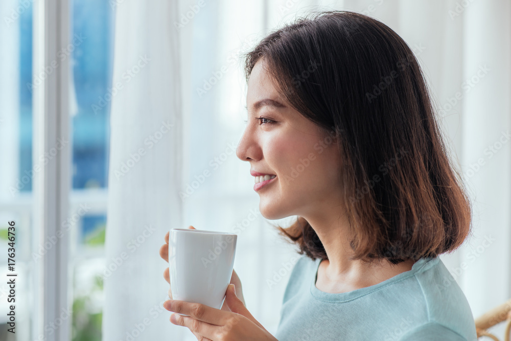 Young pretty woman sitting at opened window drinking coffee and looking outside enjoys of rest